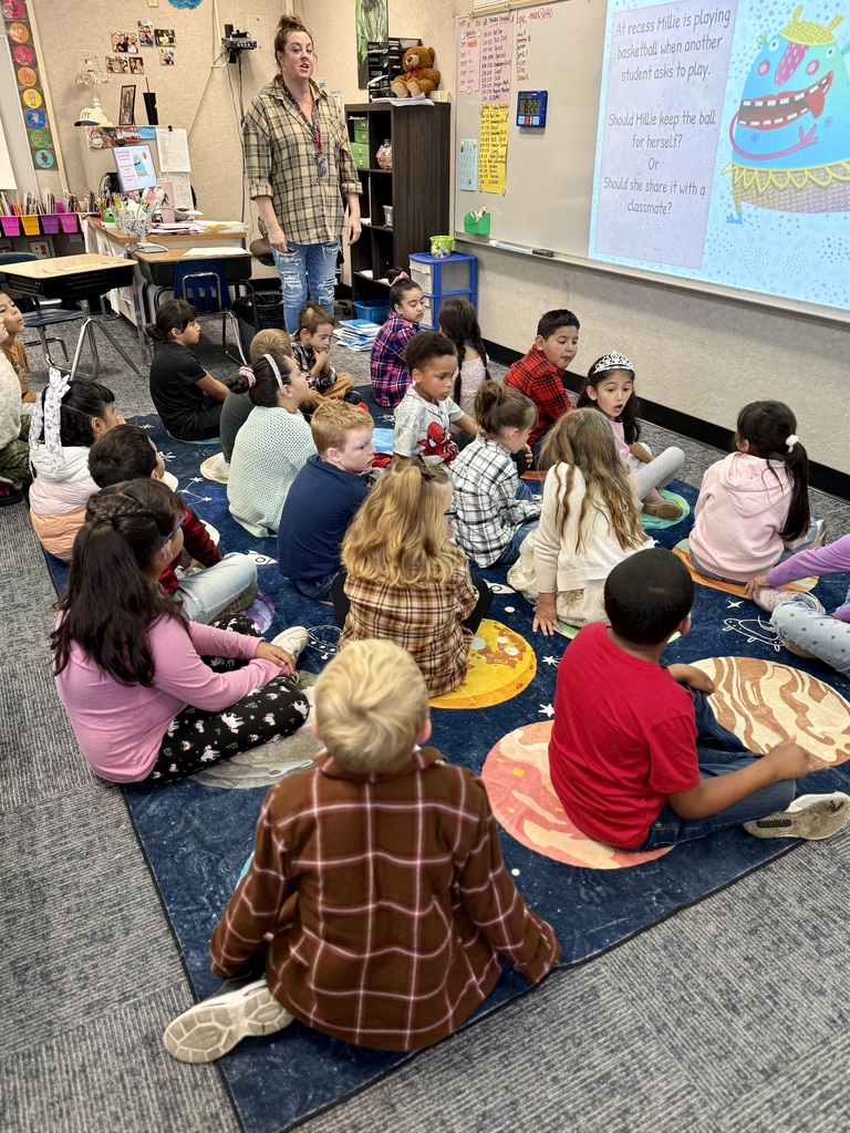 A classroom with children sitting in a circle on a rug. A teacher stands in front of a screen.