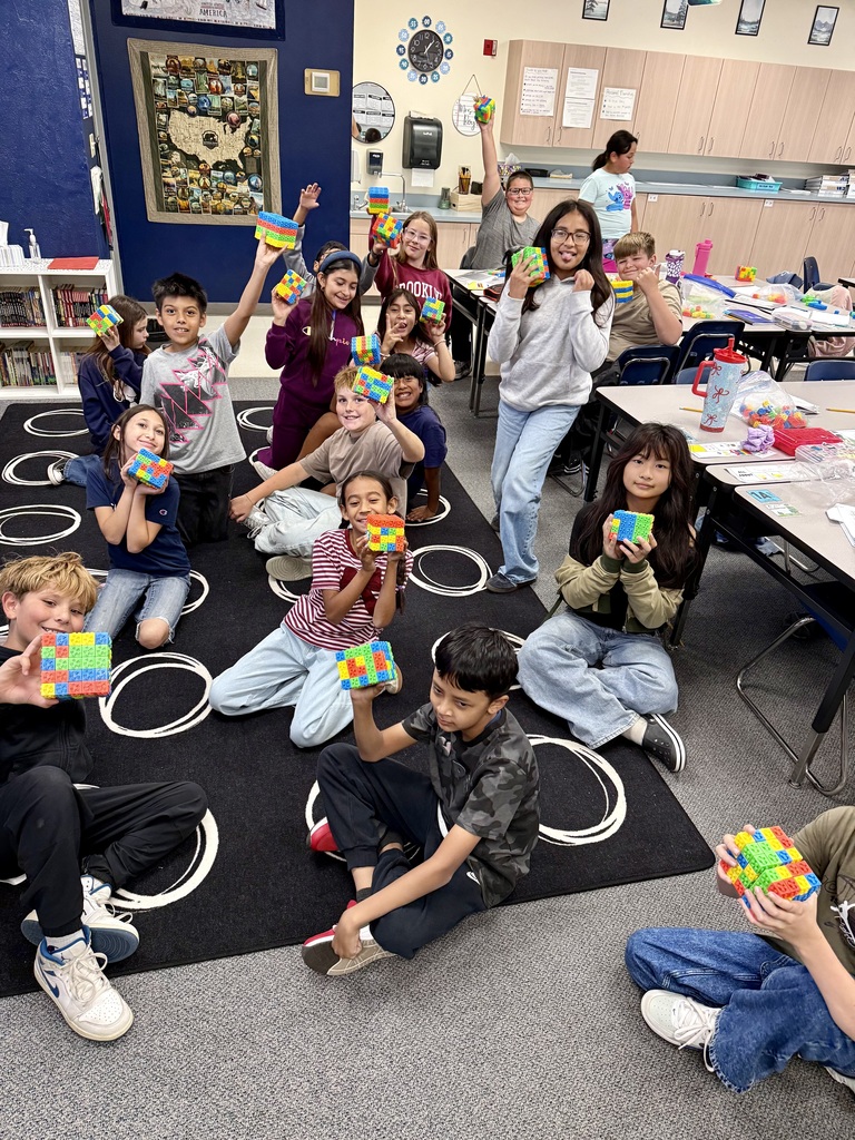 Children sit on the floor in a classroom, holding colorful cubes. A teacher stands among them.