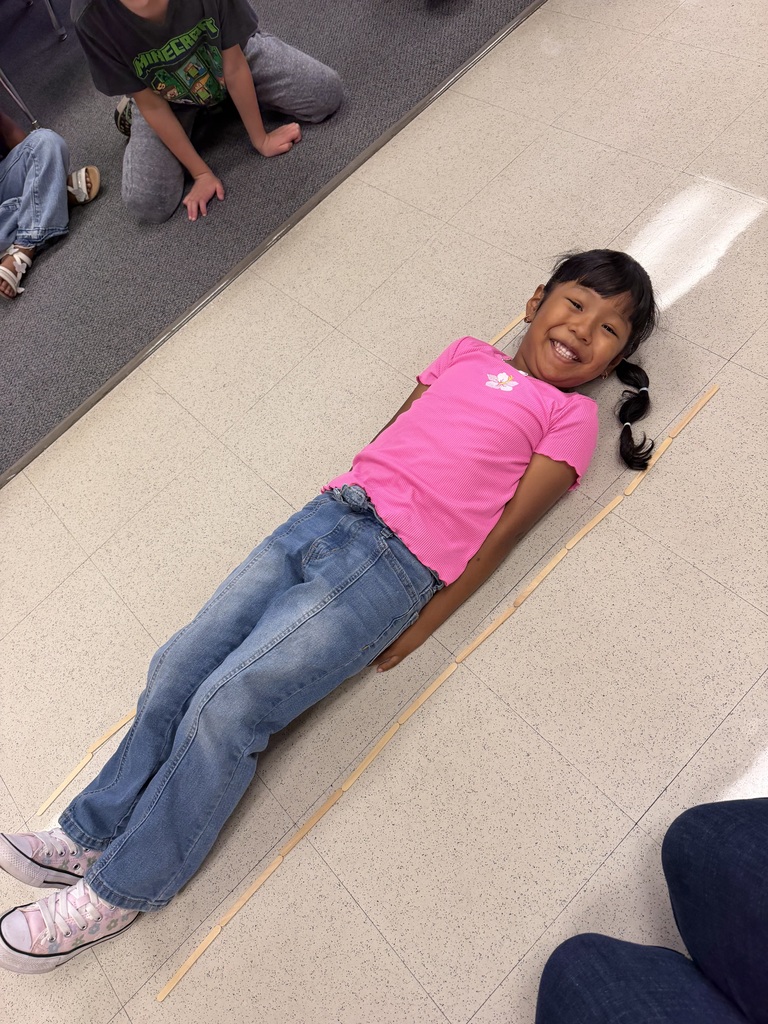 A child lies on the floor with a smile, measuring distance with sticks, in a classroom with tiled floor.