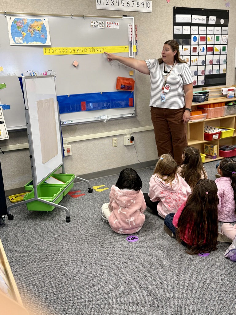 A woman stands in front of a whiteboard with a map, teaching young students sitting on the floor.