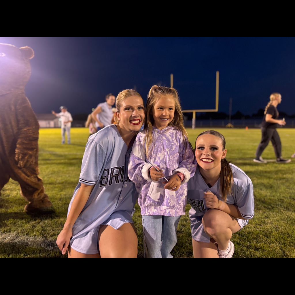Three people smile for a photo on a field at night. A mascot is behind them.