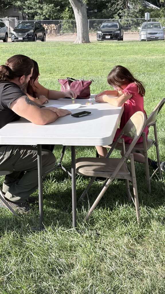 People sit at a table in a park. The table has a phone and a pink bag. Cars are parked in the background.