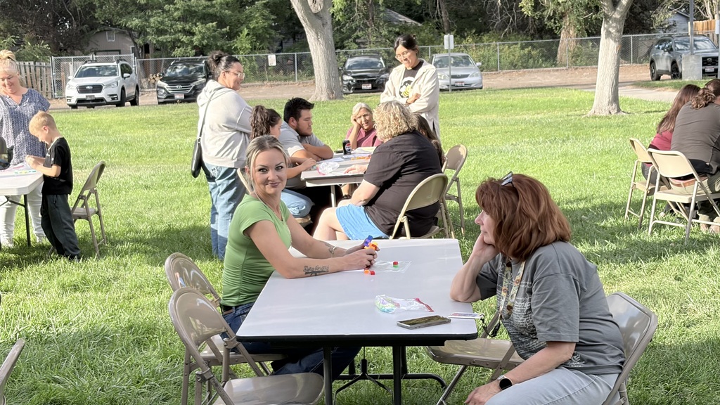 People in a park with tables and chairs, some seated and some standing, near cars parked in the background.