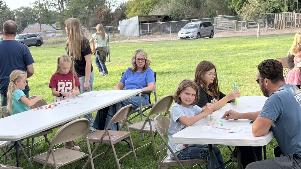 Families sit at tables outside with crafts. One person holds a paintbrush. The background shows trees and a parking lot.