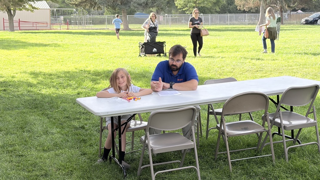 A man and a child sit at a long table on a grassy field. Behind them are people walking.