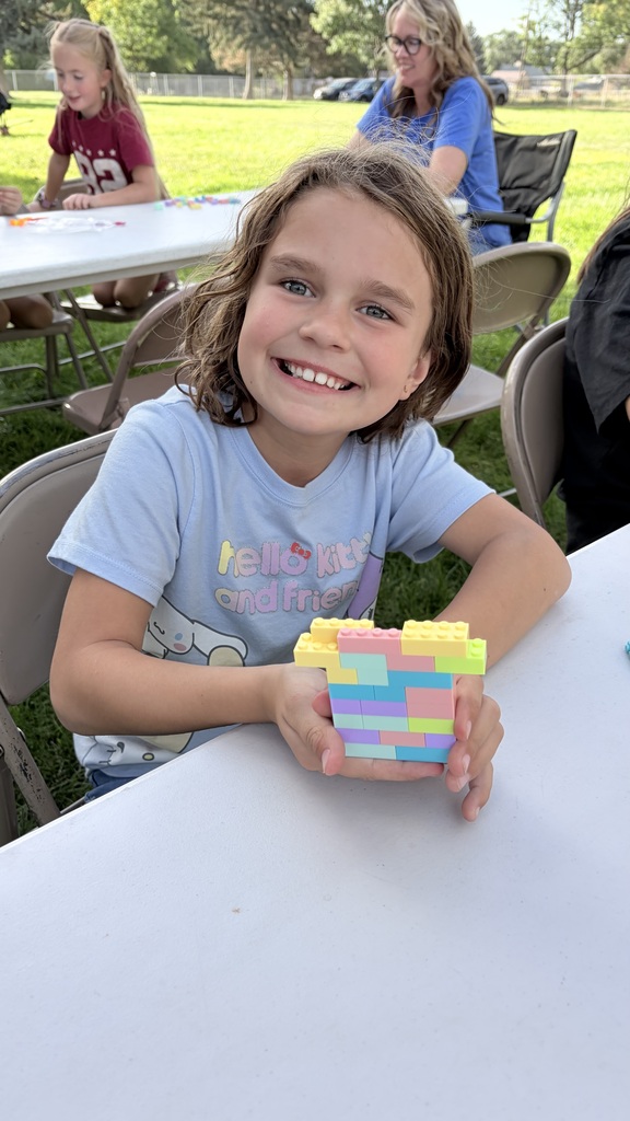 A girl wearing a blue shirt holds colorful Lego blocks, sitting at a white table. Other children are seated at tables behind her, with a green grassy background.