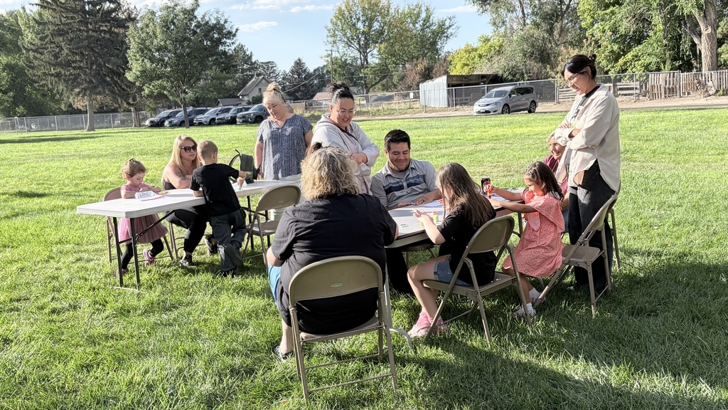 A group of people sits around tables on a grassy field. Some wear glasses, and one person stands.