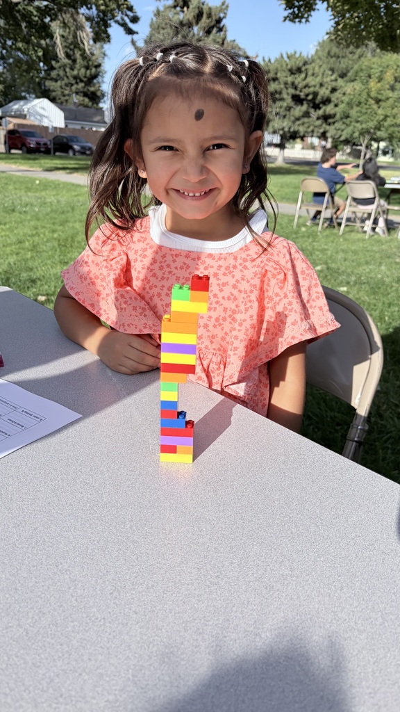 A young girl in a pink dress sits at a table, smiling, with a colorful block tower in front of her.
