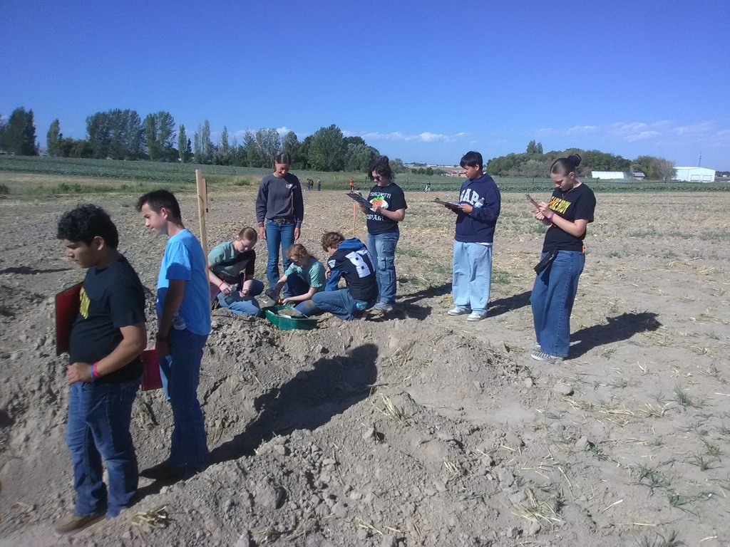 A group of people, some standing, some sitting, and some holding objects, stand in a dirt field.