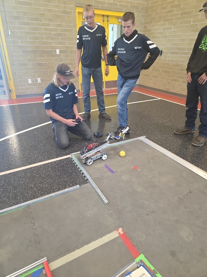 Four individuals are in a gym. One person is kneeling and holding a remote control. Another person is standing, looking at the remote.