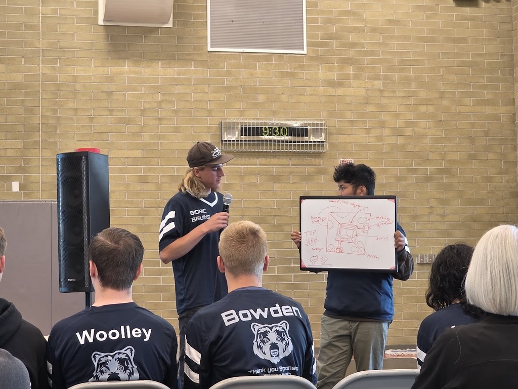 Group of people seated, two men stand before them. One man holds a microphone, other holds a whiteboard. Brick wall behind them.