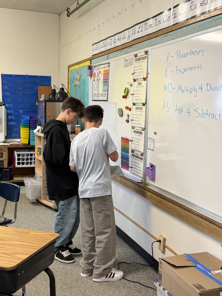 Two students in a classroom, standing and facing a whiteboard with educational content. A desk and chair are in the foreground.