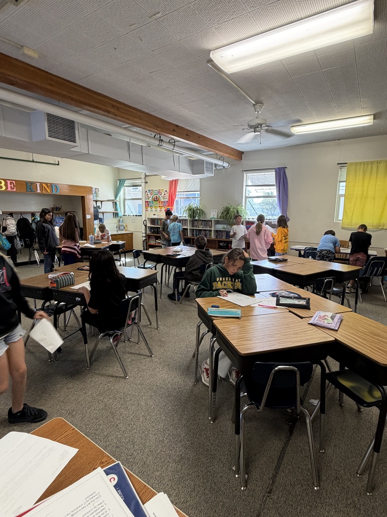 A classroom with desks and chairs arranged. Students are seated, and some are standing. Bright lighting is on the ceiling.