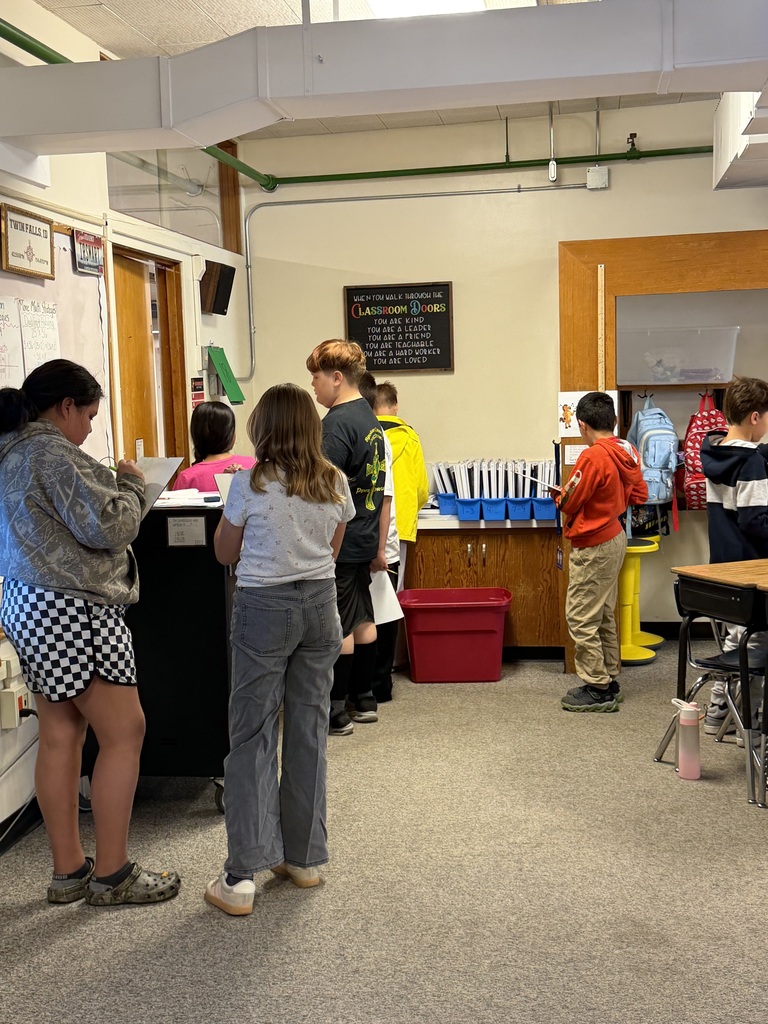 A classroom with students; some standing, others near a table. A blackboard and bulletin board are on the wall.
