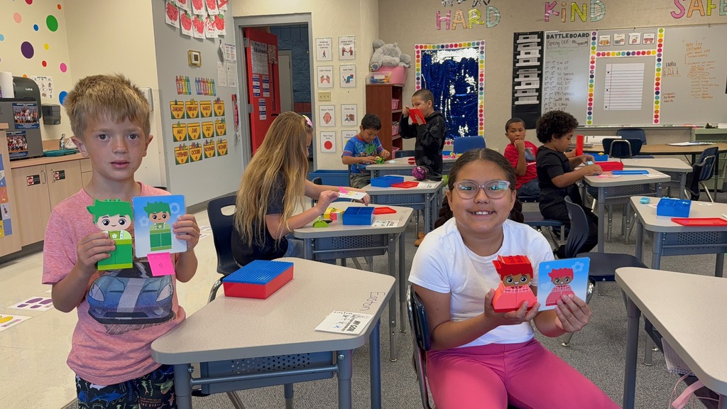 Kids in a classroom, one boy with red shorts holds a toy while another girl in pink shorts holds a doll.