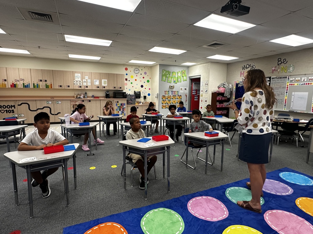 Classroom with students seated at desks, a woman standing in front, and colorful floor mats.