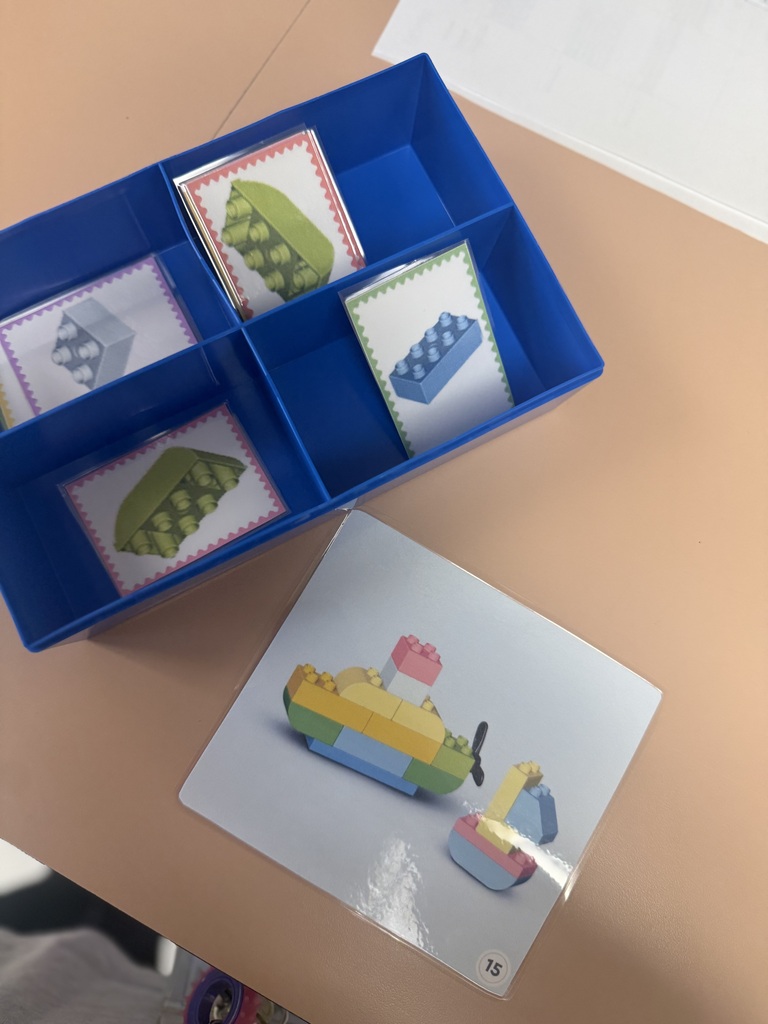 A blue tray with several compartments on a table, each containing small, colorful Lego-like pieces. A larger card with a Lego creation image is next to the tray.
