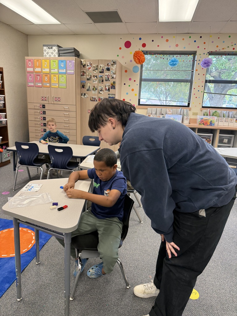 A classroom with desks and chairs. A person leans over a desk with a child seated.