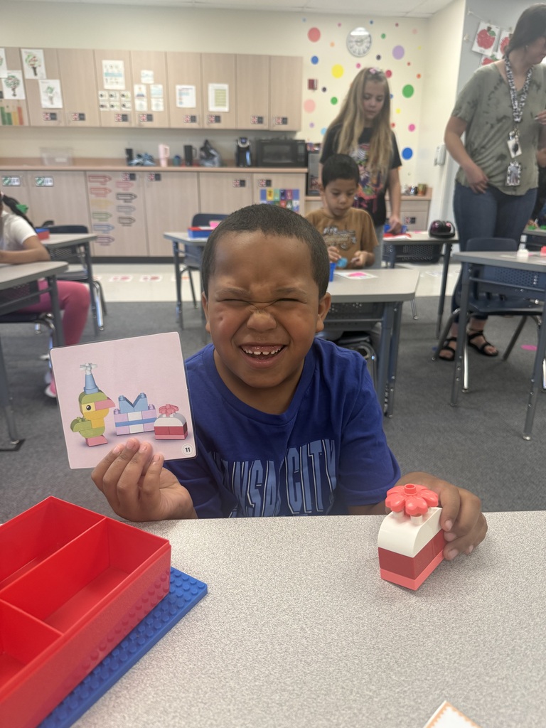 A young boy in a blue shirt holds a card with a drawing. He sits at a desk in a classroom with other students.
