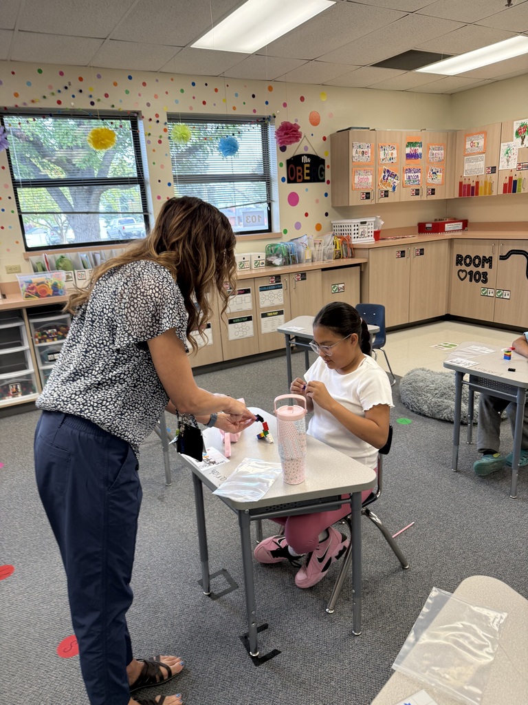 Teacher assisting a student at a desk in a classroom with a colorful wall and a carpeted floor.