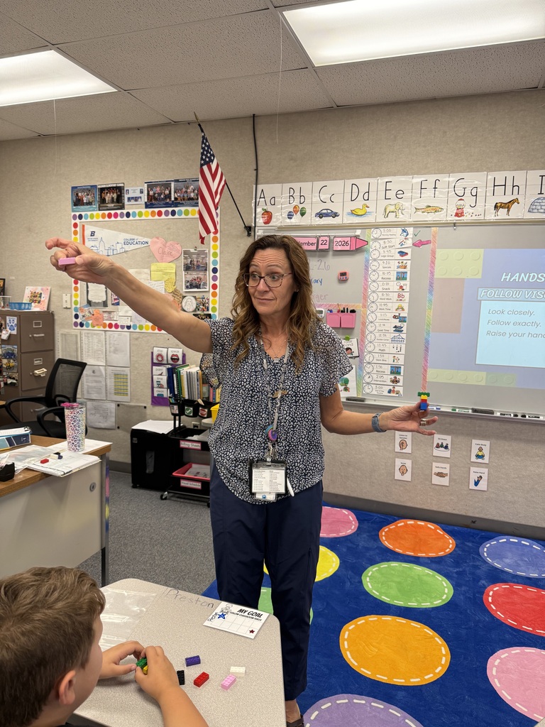 A teacher demonstrates with hands in a classroom with a boy sitting at a desk.