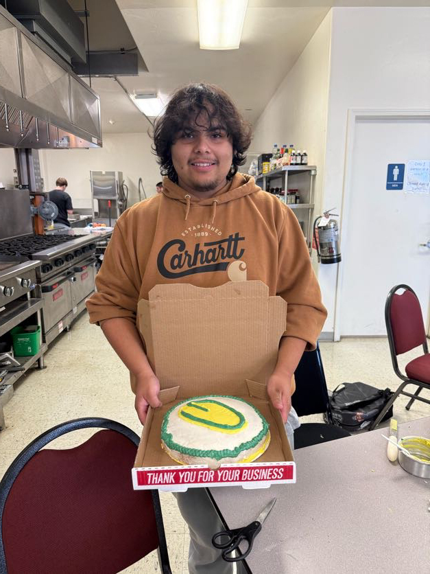 Person in a kitchen with a box of cake, smiling. Behind them is a stove and shelves.