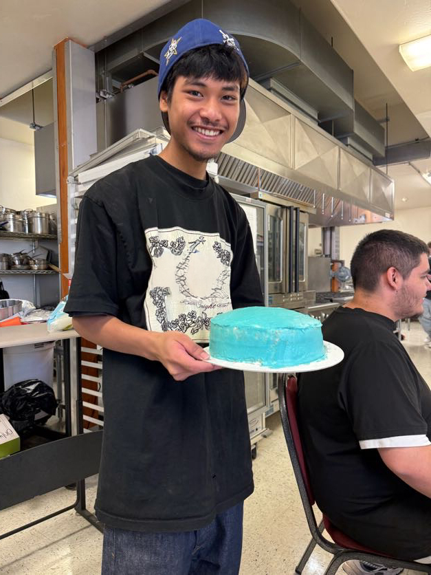Person holding a plate with a blue cake in a kitchen. A man sits in a chair behind.