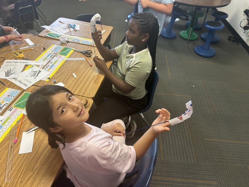 Two girls, one with a paper airplane, sit at a desk with other students in a classroom.