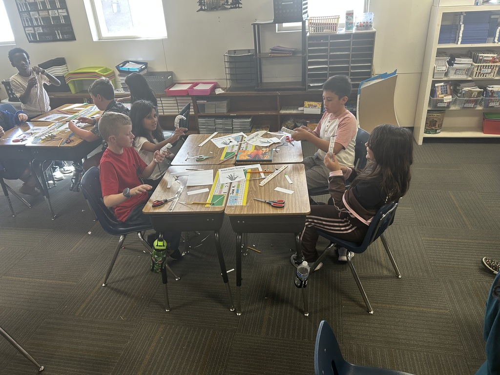 Several children work at tables in a classroom, with paper, scissors, and other items. Bookshelves in background.