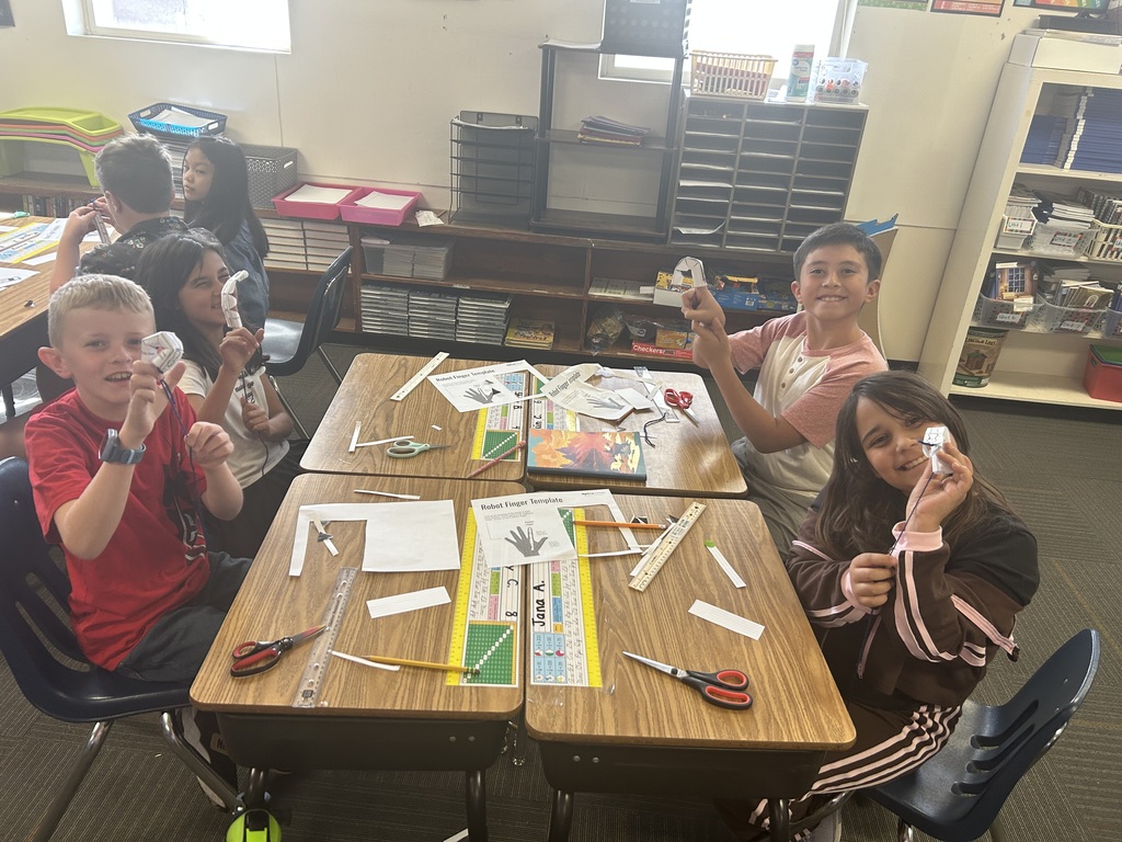 Children seated at desks, holding scissors and paper. Two desks hold craft materials, including paper and scissors. A shelf with books is behind them.