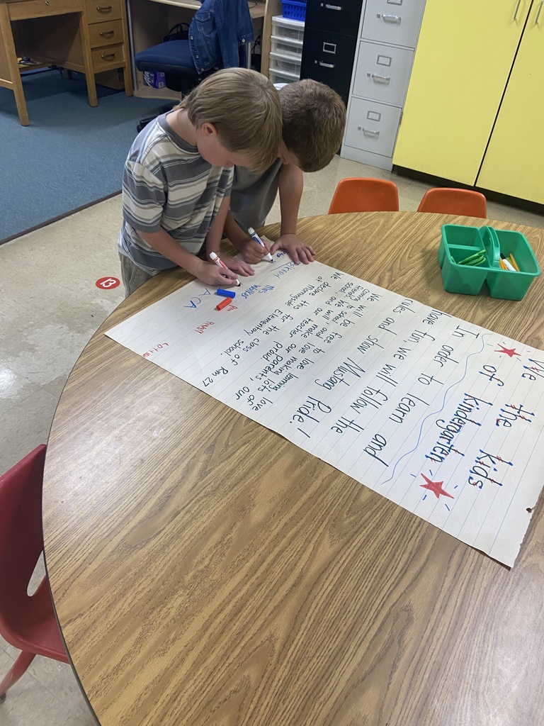 Two children are writing on a large sheet of paper on a table in a classroom.