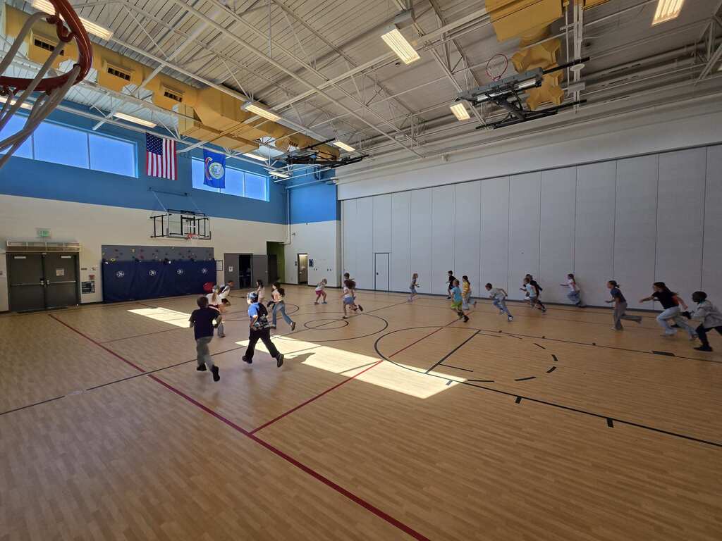 An indoor basketball court with people playing. The court has a wooden floor, red lines, and a flag on the wall.
