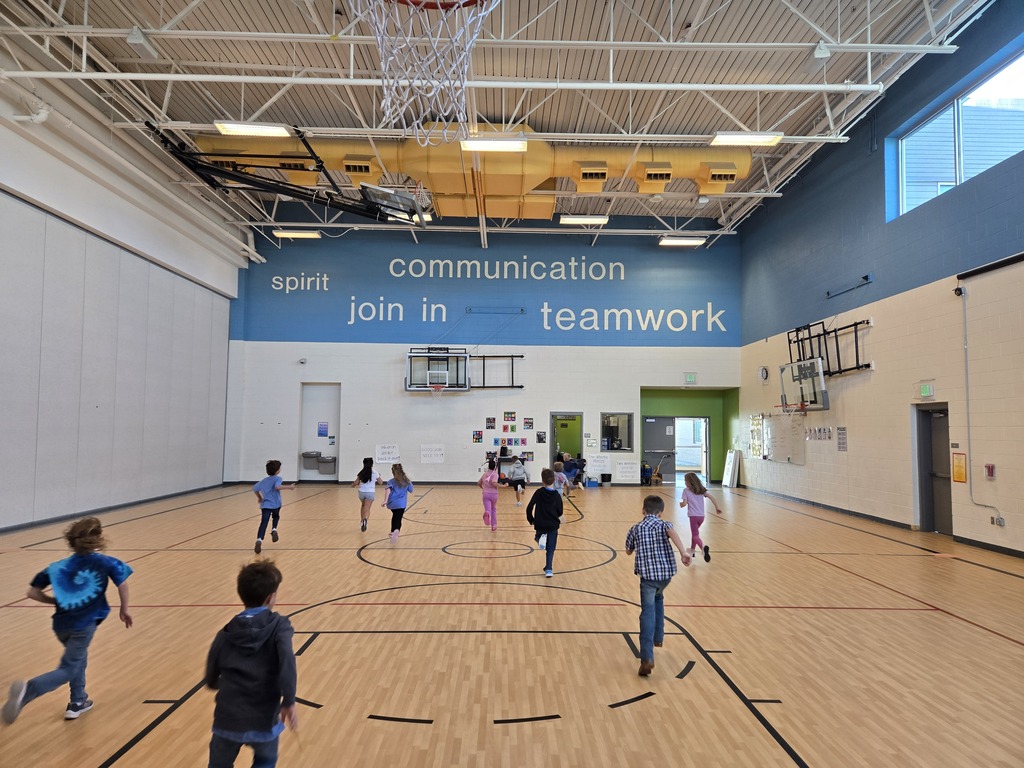 Children run and play on an indoor basketball court. Walls are white and blue, with words like "communication" and "teamwork" on the wall.