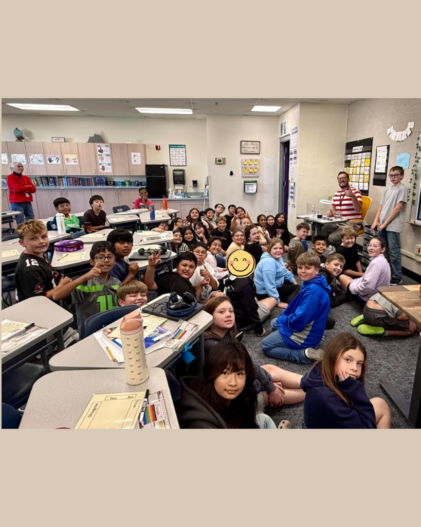 A classroom with desks, students seated and standing, one person smiling at the camera, a teacher near the door, and various items on desks.