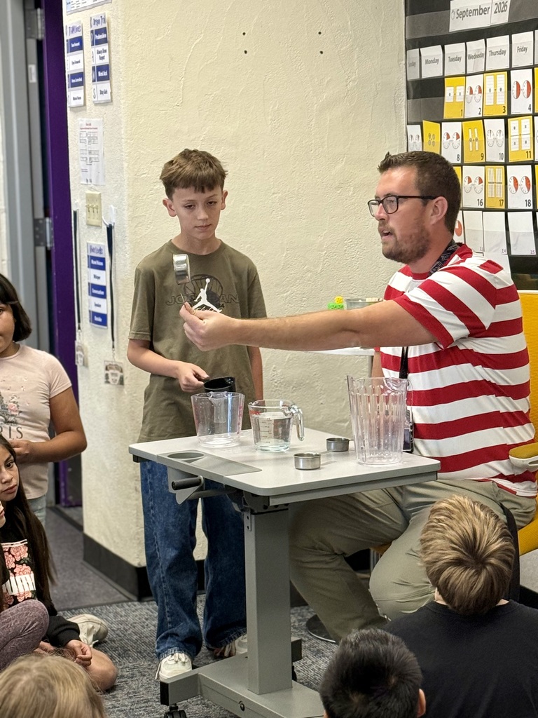 Man seated at a table demonstrating a science experiment with a boy. Students seated and watching.
