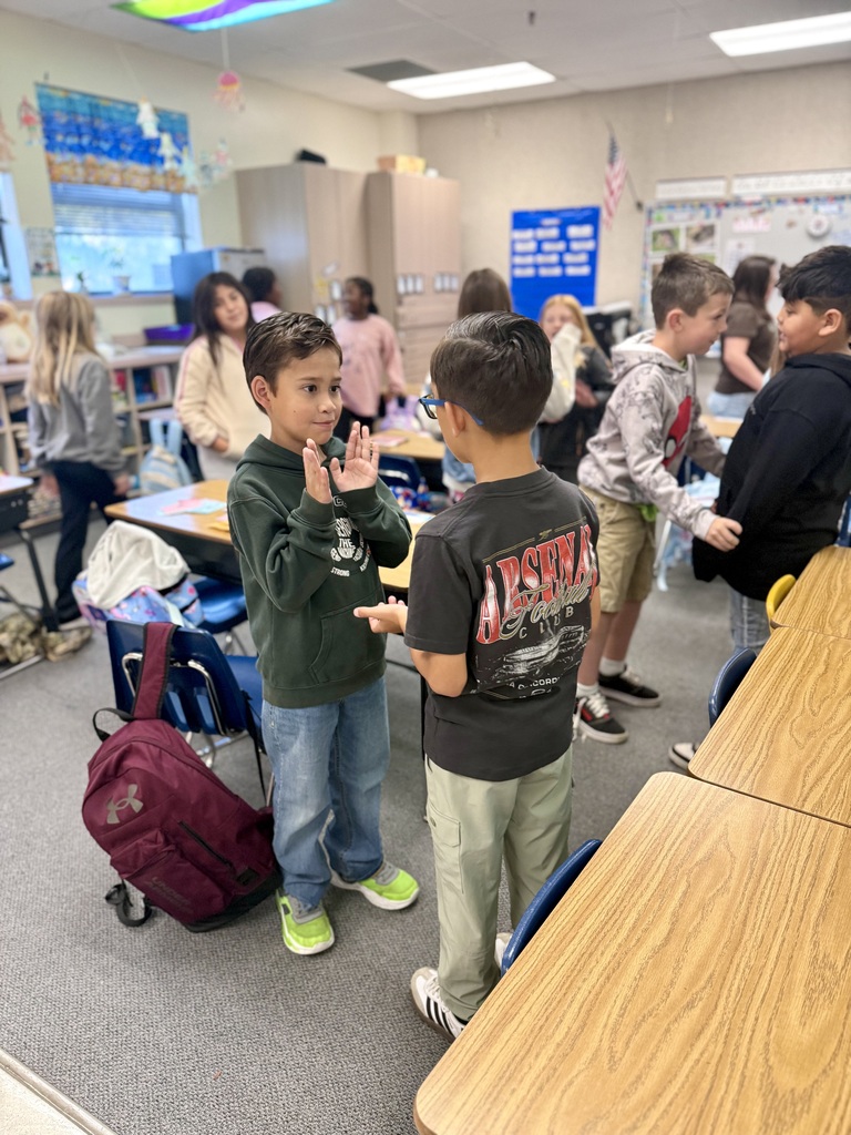 A classroom with children standing and talking. One boy uses sign language. A table has a red backpack.