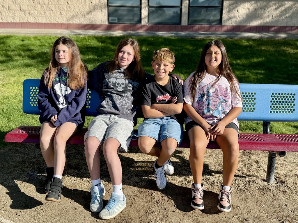 Four children pose for a photo on a bench, two girls in front and two boys behind them. A building with glass windows is in the background.