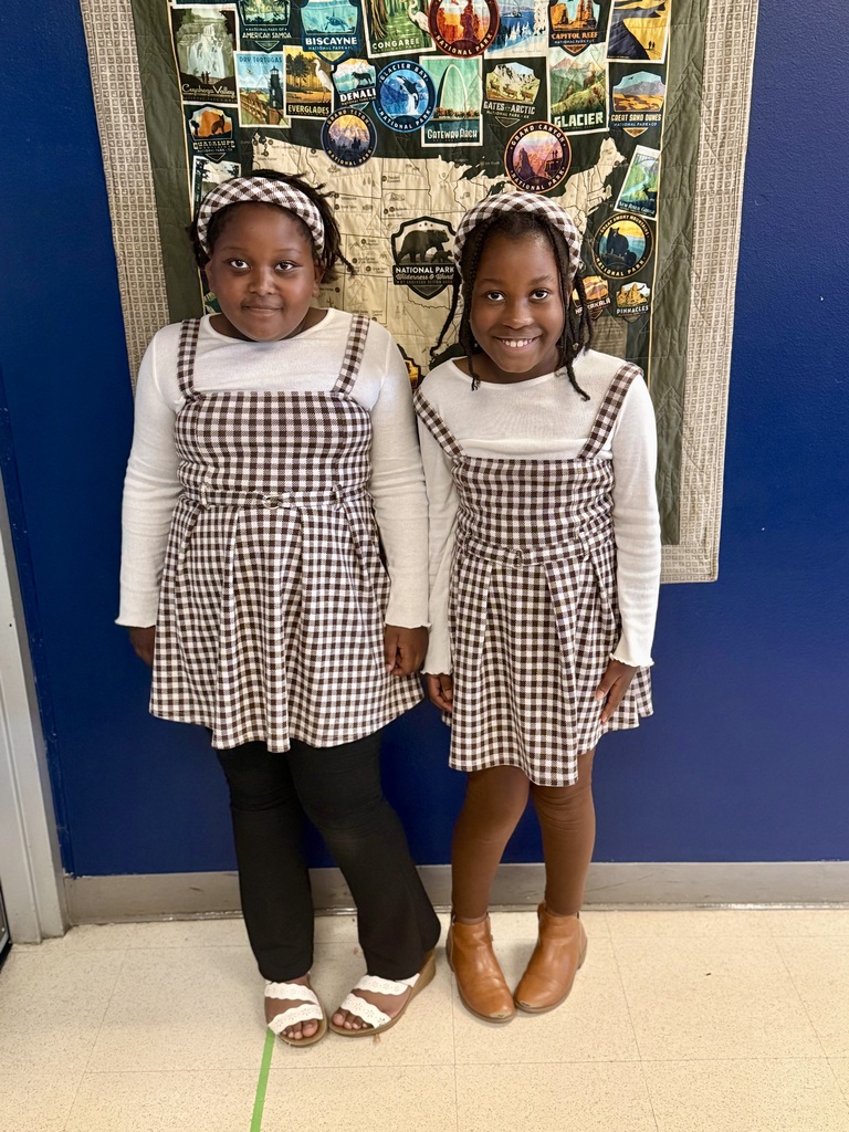 Two girls in checkered dresses, one in black pants and the other in brown boots, stand in front of a map.