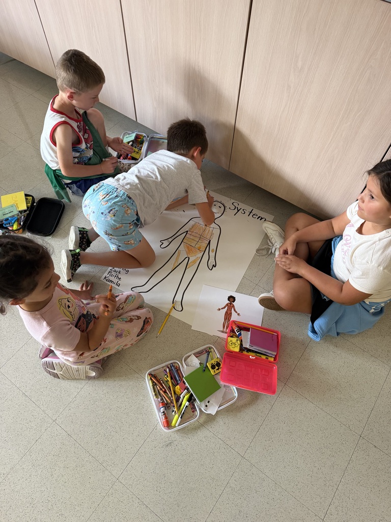Four children are seated on the floor, engaged in a creative activity. They have art materials and a large diagram of the human body spread out.
