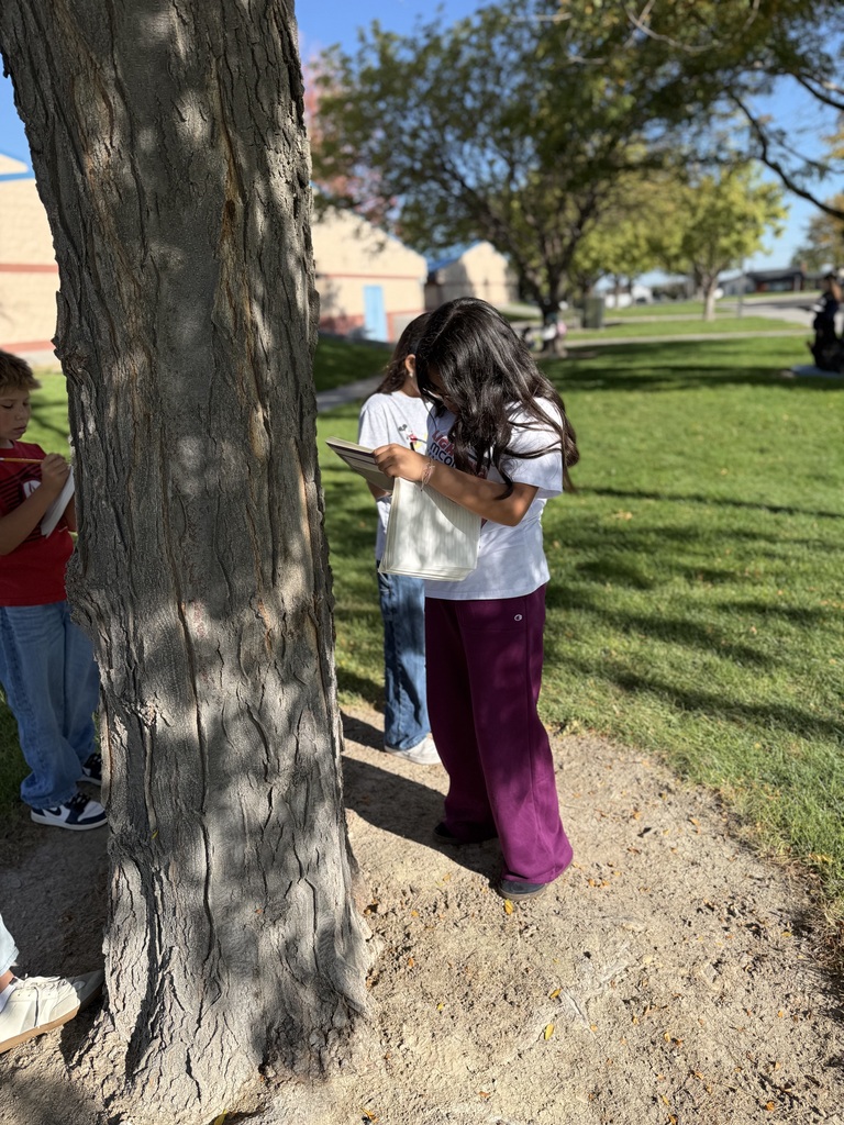 Three children are standing by a tree. One holds a tablet. Green grass and buildings in the background.