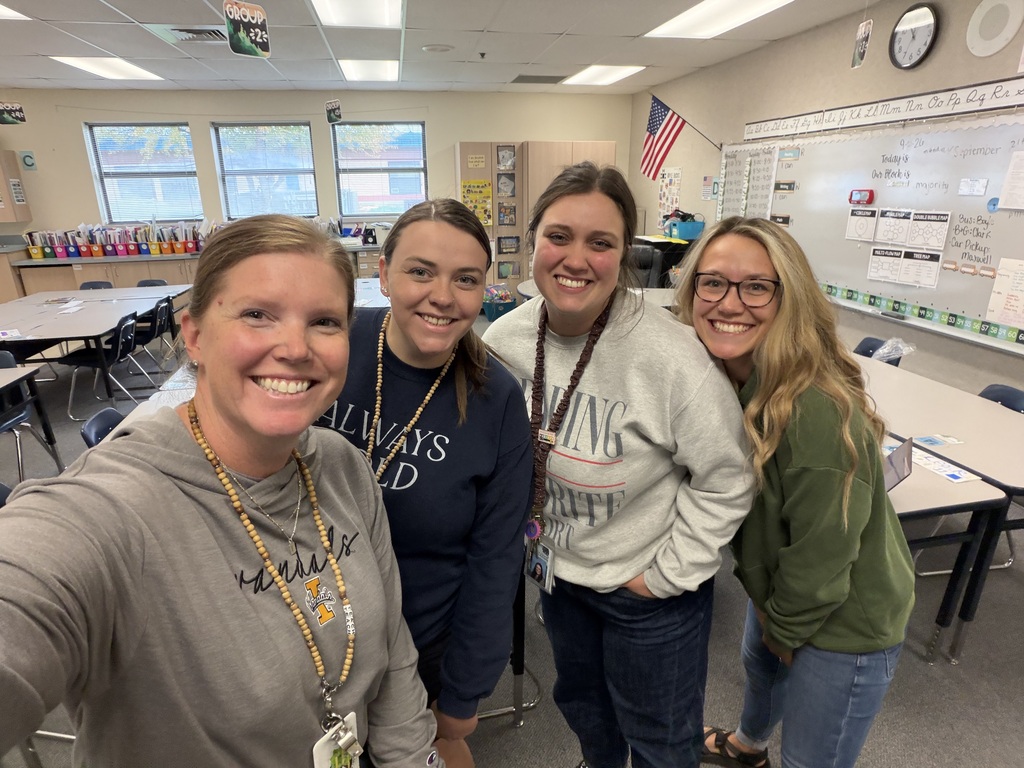 Four women in a classroom. They wear casual clothes and are smiling. A table and chairs are in the background.