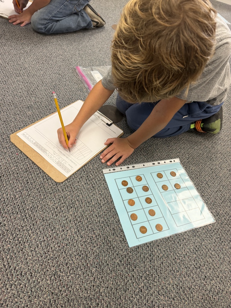 A child sits on the floor, using a pencil to write on a paper next to a grid of coins.