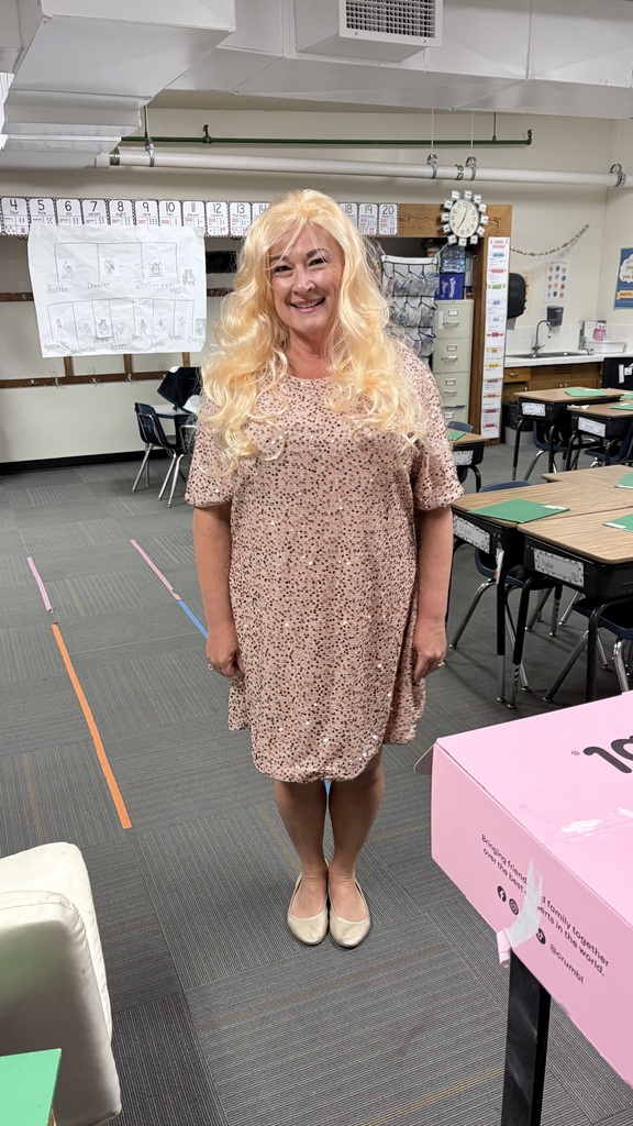 A woman with blonde curly hair, smiling, stands in a classroom with tables and chairs in the background.
