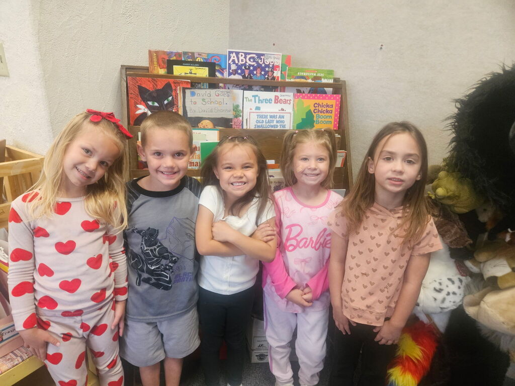 Five young girls in pajamas stand together in a room with a white wall, shelves with books, and toys.