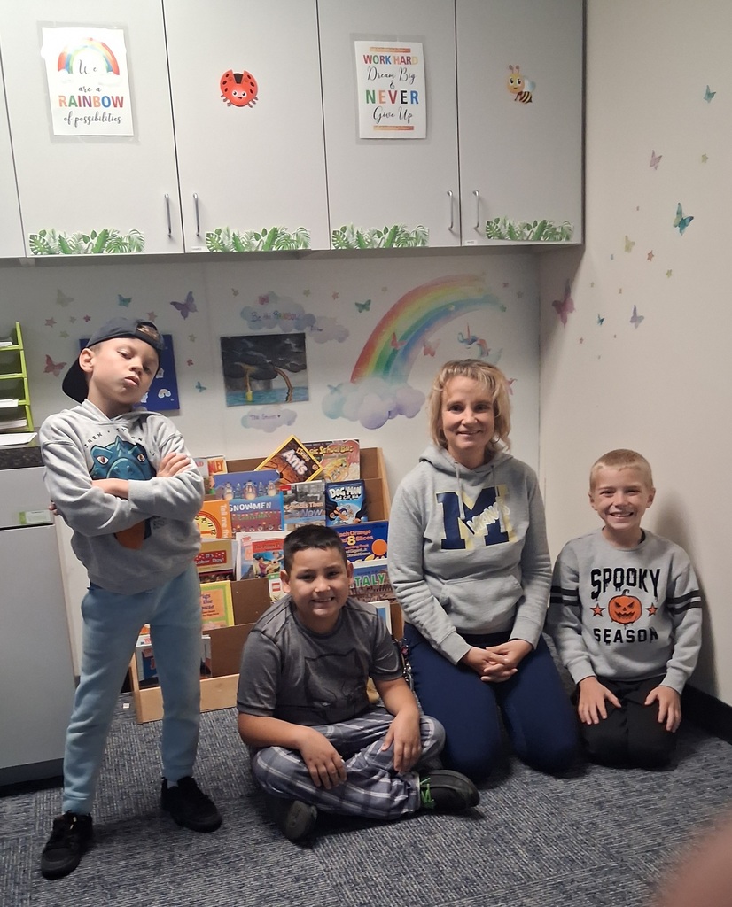 Four children pose for a photo in a classroom. Behind them is a wall with colorful decorations and a rainbow.