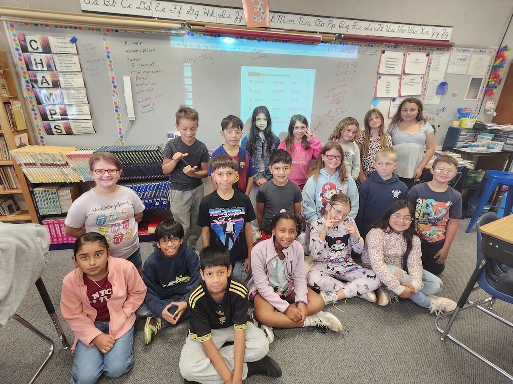 Children in a classroom, some seated, some standing, in front of a white board and shelves.