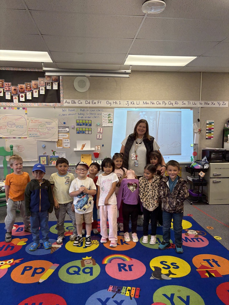 A group of children in a classroom, standing on a colorful alphabet rug. A woman is in front.