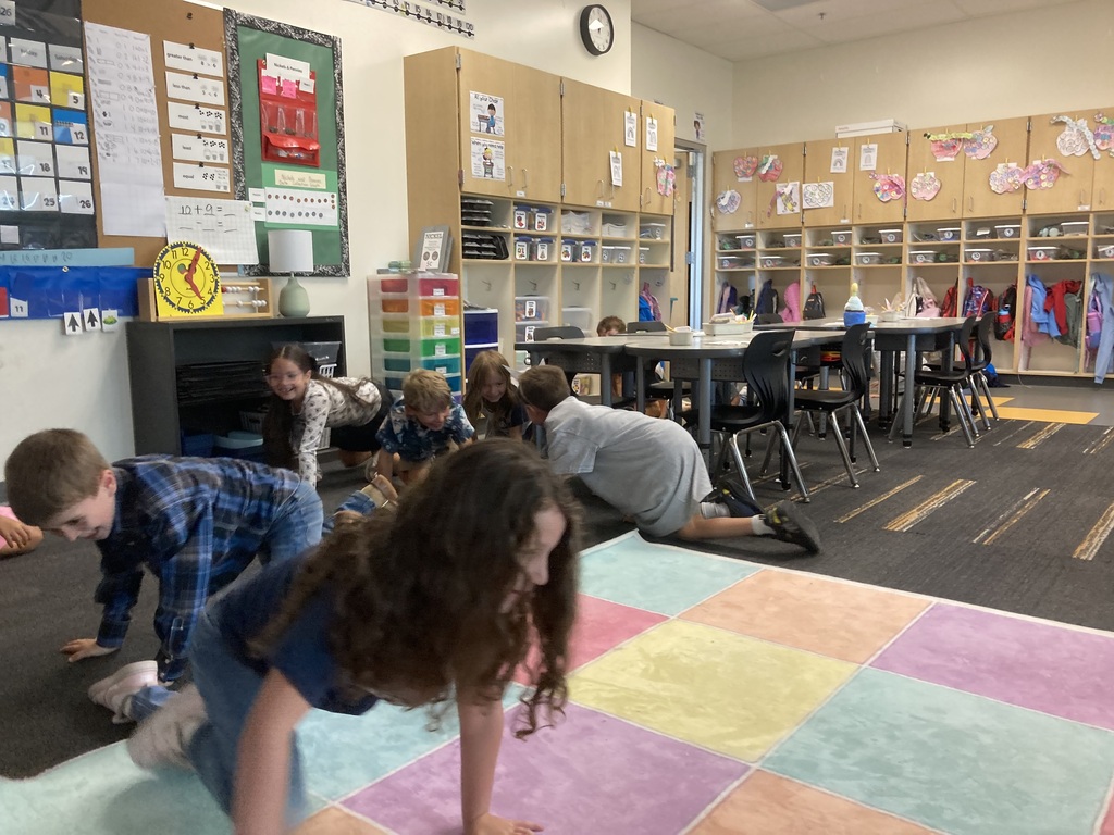 A classroom with children doing floor exercises on a multicolored mat. Shelves with items and tables with chairs are in the background.
