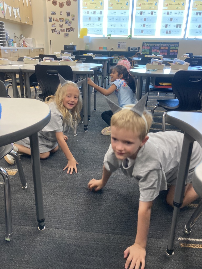 Kids in a classroom wearing small pointed hats. A boy crawls on the floor while a girl crouches nearby.