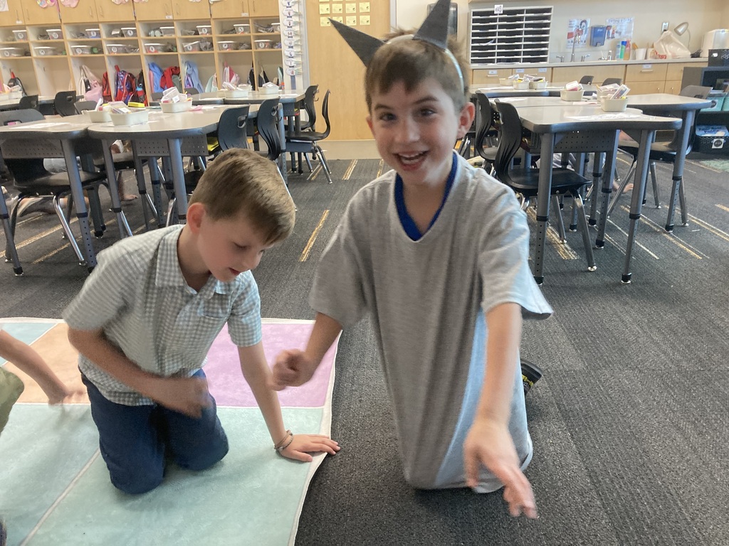 Two boys, one kneeling and one standing, engage in an activity in a classroom with tables and chairs.